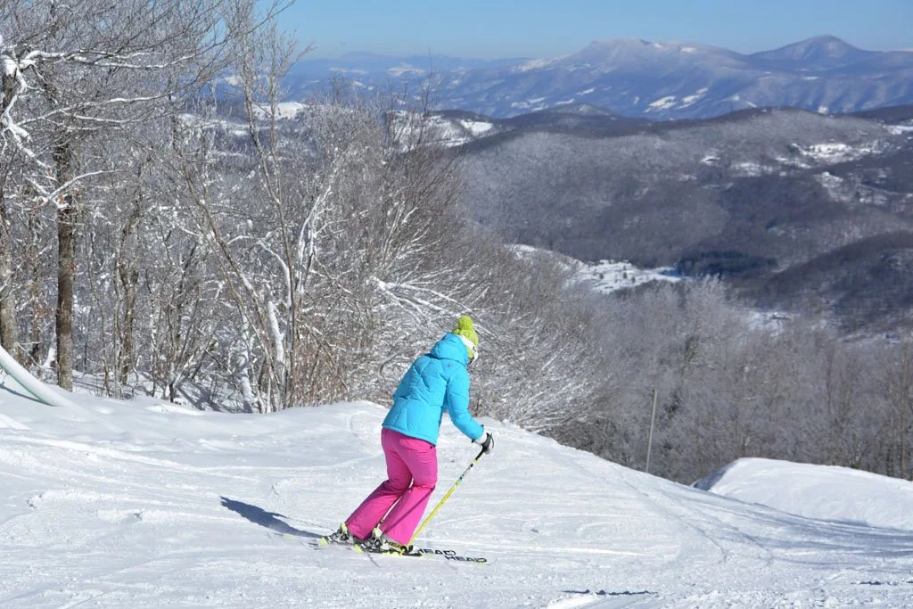 A skier descending a snowy Sugar Mountain slope with Blue Ridge peaks beyond